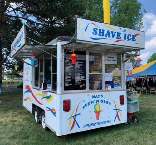 A colorful shaved ice stand with a menu, located outdoors under a blue sky and surrounded by trees.
