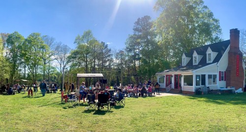 A sunny outdoor gathering with people socializing near a house, surrounded by trees and lawn chairs.