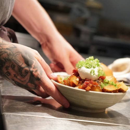 A close-up of a hand with a tattoo, serving a bowl of food topped with bacon, green onions, and a poached egg.