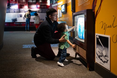 A man and a child point at an old television in a museum, surrounded by colorful exhibits.