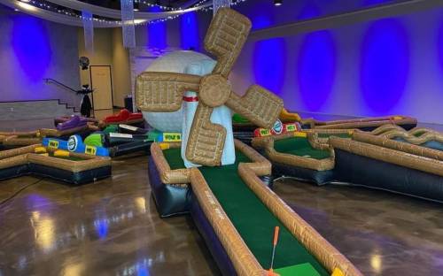 A colorful indoor mini-golf course featuring inflatable obstacles, including a large windmill, under purple lighting.