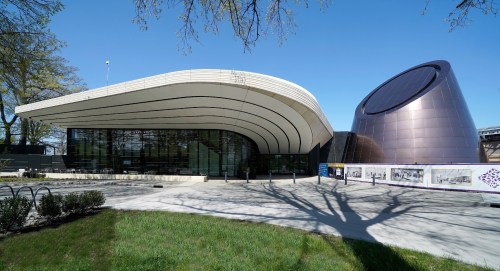 Modern architectural building with a curved roof and a metallic dome, surrounded by green grass and trees.