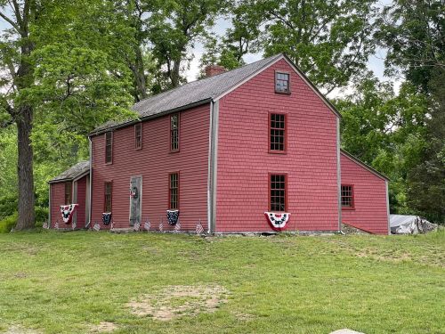 A red, two-story house with decorative bunting, surrounded by greenery and trees on a cloudy day.
