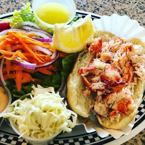 A lobster roll on a bun with a side salad, coleslaw, lemon wedge, and a small cup of dressing.