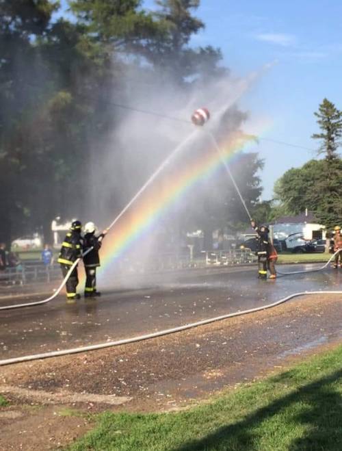 Two firefighters spray water, creating a rainbow in the mist, with trees and a clear sky in the background.