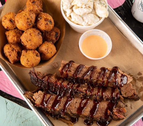 A tray featuring sliced brisket with barbecue sauce, a side of coleslaw, and golden cornbread muffins.