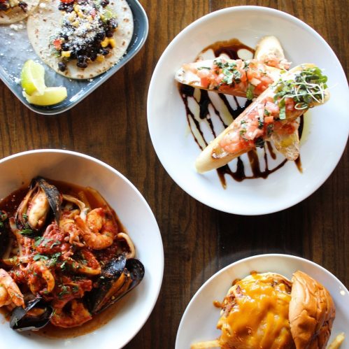 A top-down view of a table with various dishes: tacos, shrimp, seafood pasta, and a cheeseburger with fries.