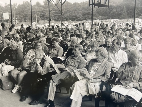 A large crowd of elderly people seated under a pavilion, some reading, others engaged in conversation.