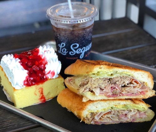 A plate with a sandwich filled with meats, a slice of cake topped with whipped cream and cherries, and a drink.