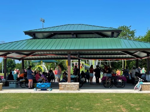 A large group of people gathers under a green-roofed pavilion in a park on a sunny day.