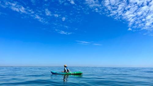 A person in a kayak paddles on calm water under a clear blue sky with scattered clouds.