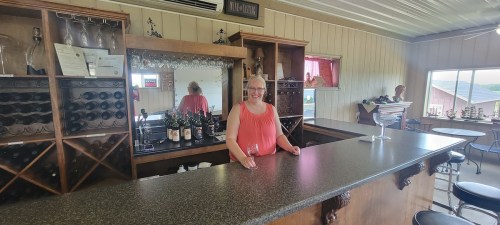 A woman stands at a bar with wine bottles behind her, smiling and holding a glass in a cozy, well-lit space.