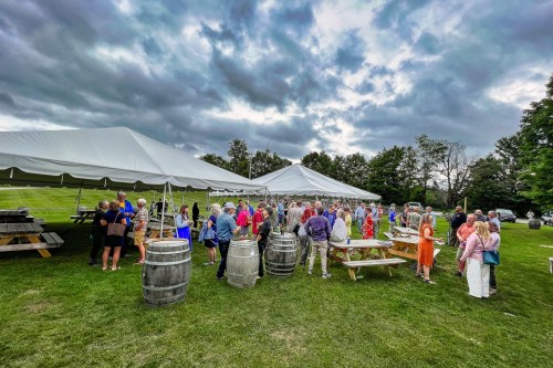A large outdoor gathering under tents, with people mingling around wooden barrels and picnic tables on a cloudy day.