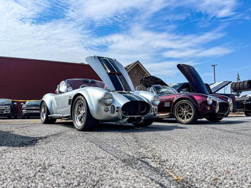 Two classic sports cars parked with hoods open, showcasing their engines, under a blue sky with scattered clouds.