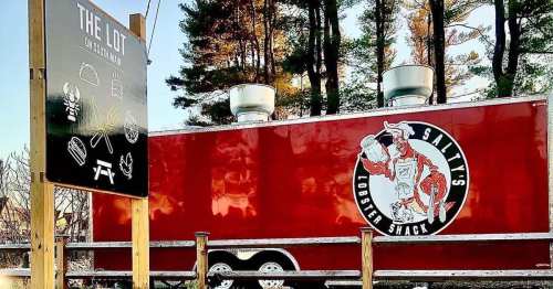 A red food truck with a lobster logo, parked near a sign for "The Lot" featuring food icons. Trees in the background.