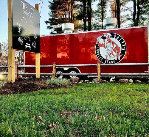 A red food truck with a lobster logo parked near a sign for "The Lot," surrounded by grass and trees.