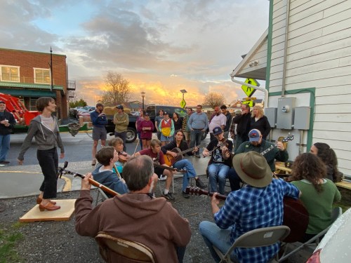 A group of people playing guitars and singing together outdoors, with an audience gathered around them at sunset.