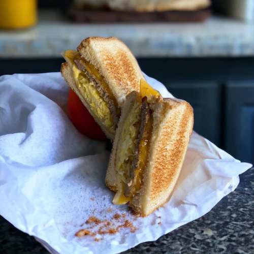 A toasted sandwich with melted cheese and egg, cut in half, resting on white paper with a tomato in the background.