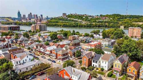 Aerial view of a riverfront city with houses in the foreground and a skyline in the background, surrounded by greenery.