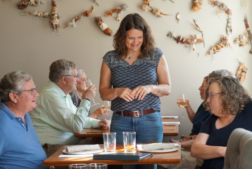A woman stands at a table in a restaurant, engaging with diners while others enjoy drinks in the background.
