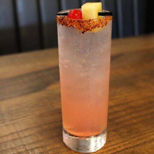 A tall glass of pink cocktail garnished with a cherry and pineapple, rimmed with chili powder, on a wooden table.