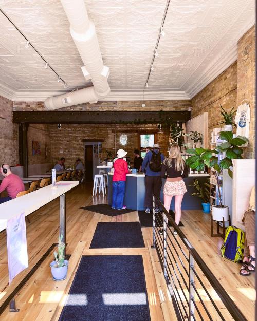 A cozy café interior with patrons at the counter, plants, and wooden floors, featuring a bright and inviting atmosphere.