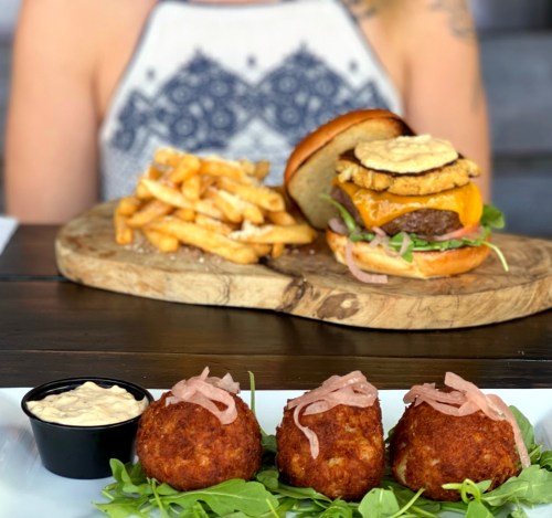 A gourmet burger with fries on a wooden board, alongside three crispy balls with sauce and garnished with onions.