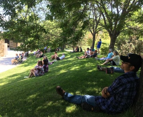 A group of people sitting on a grassy hill under trees, enjoying a sunny day outdoors.