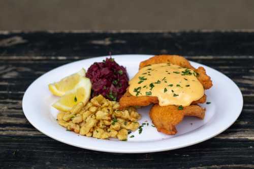 A plate of fried fish topped with creamy sauce, served with lemon, red cabbage, and a side of crispy fried dough.