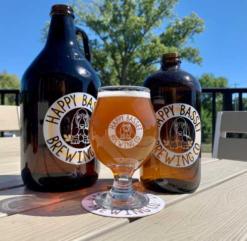 A glass of amber beer sits on a table, flanked by two growlers from Happy Basset Brewing Co. under a clear blue sky.