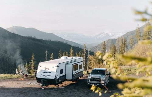 A camper trailer parked beside a truck in a scenic mountain landscape with trees and distant snow-capped peaks.