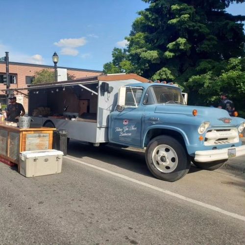 A vintage blue food truck parked on the street, with a vendor preparing food outside. Trees and buildings in the background.