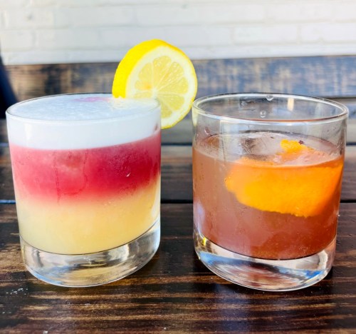 Two colorful cocktails on a wooden table: one layered with yellow, red, and white, and the other brown with an orange slice.