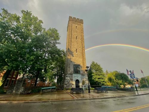 A tall stone tower stands beside trees, with a rainbow arching across a cloudy sky after rain.