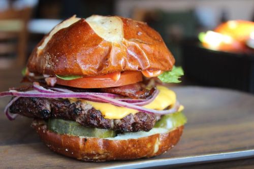 A delicious burger with a pretzel bun, beef patty, cheese, bacon, lettuce, tomato, red onion, and pickles.
