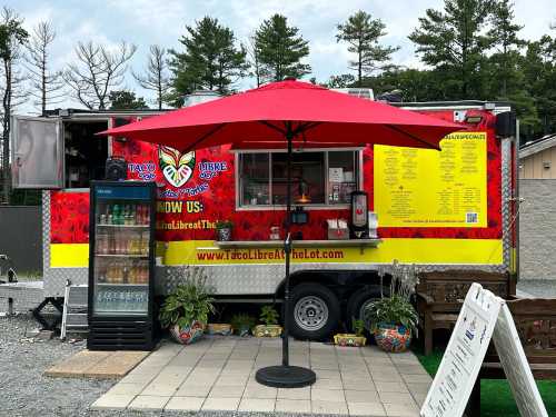 A colorful food truck with a red umbrella, featuring a menu and drinks, set in a gravel lot surrounded by trees.