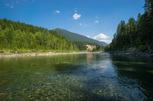 A serene river flows through a lush green landscape, surrounded by mountains and clear blue skies.