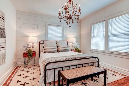 Cozy bedroom with a metal bed frame, chandelier, and natural light from two windows, featuring a rug and decorative plants.