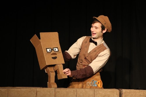 A performer in a brown outfit holds a cardboard puppet with glasses, smiling on stage against a dark backdrop.