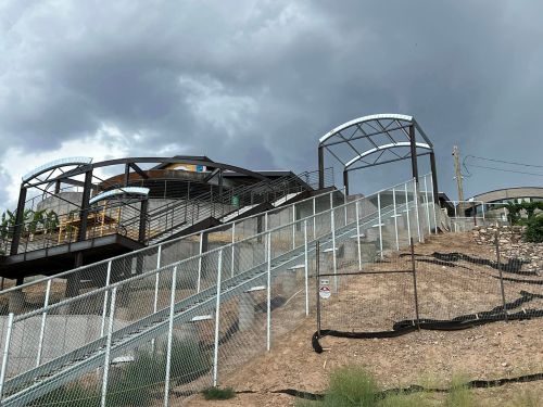 A modern building with a staircase and archways, set against a cloudy sky and surrounded by a fence.