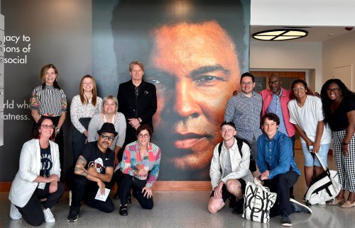 A diverse group of people poses in front of a large mural of Muhammad Ali, smiling and engaging with each other.