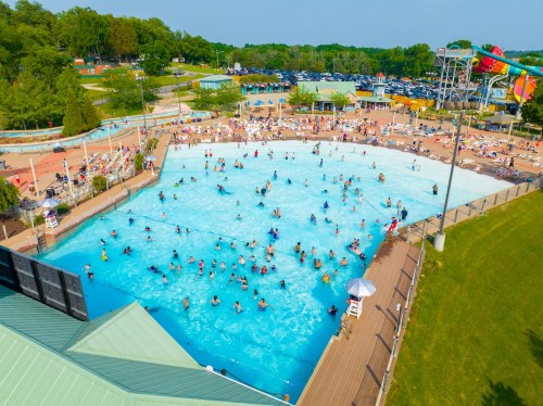 Aerial view of a crowded water park pool with people swimming, sunbathing, and water slides in the background.