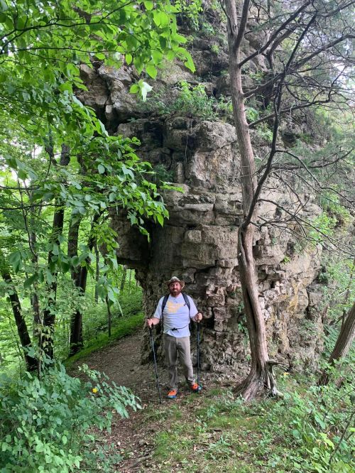 A hiker stands near a large rock formation surrounded by lush greenery on a forest trail.