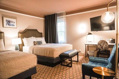 Cozy hotel room with two beds, a chair, desk, and a TV, featuring warm colors and soft lighting.