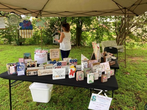 A vendor sets up a table with handmade crafts and cookies under a tent in a grassy outdoor area.
