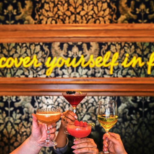 Four hands holding colorful cocktails in front of a decorative wall with the phrase "Discover yourself in fun."