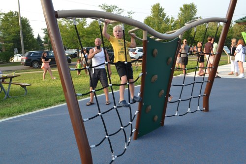 A child climbs a playground structure while an adult watches nearby, with people enjoying the park in the background.