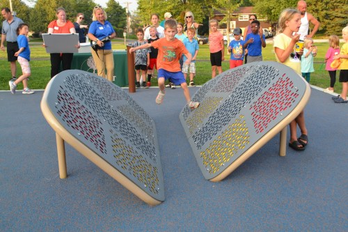 A boy jumps between two large, colorful playground structures while other children and adults watch nearby.