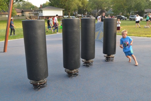 A child runs past four large, cylindrical spring structures on a playground, with people enjoying the park in the background.