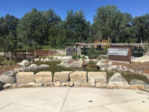 A serene park scene featuring a pond, rocks, and a sign for Foster Waterfowl Refuge, surrounded by trees and greenery.